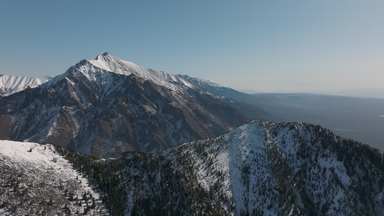 Ampie riprese aeree del Picco Galina innevato e di aspre creste montuose sopra il villaggio di Arshan, Repubblica di Buriazia, Russia, con cielo sereno e orizzonte lontano.