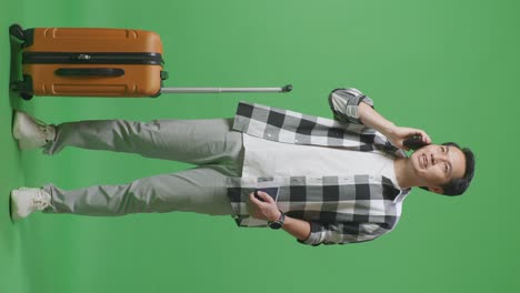 full body of asian male traveler with luggage and passport talking on smartphone while standing in the green screen background studio