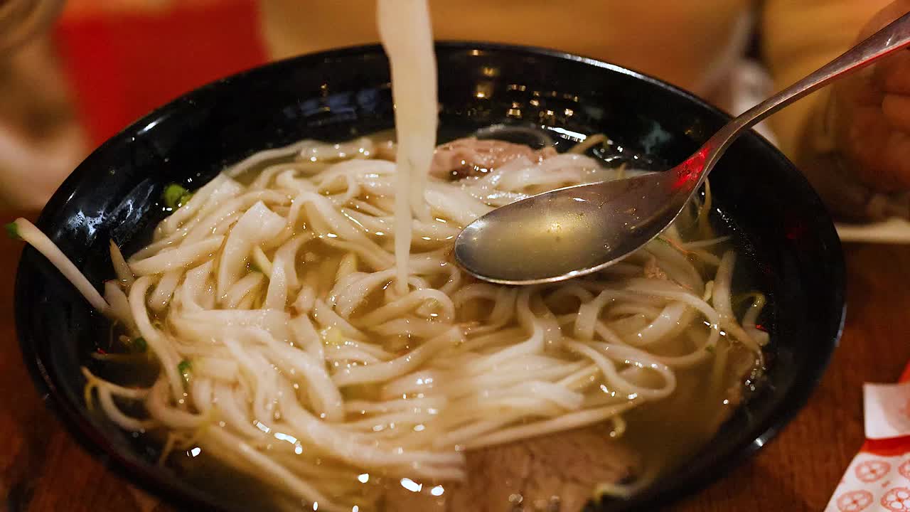 A person savors a bowl of Vietnamese pho using chopsticks and a spoon in a warmly lit Gold Coast setting