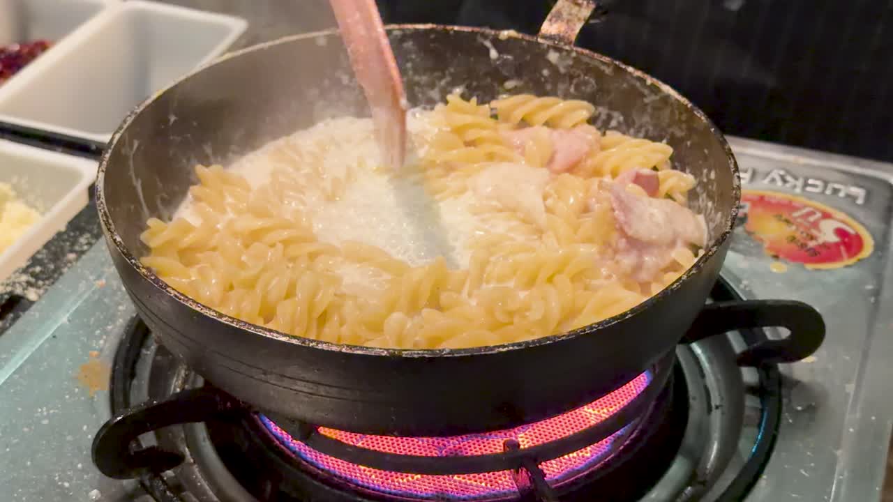 Fusilli pasta and bacon stirred in creamy sauce over open flame, close-up, warm lighting