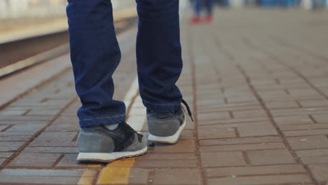 young boy in jeans and trainers is waiting for express train on railway station platform. boy's legs going forward and backward on the pavement. close-up
