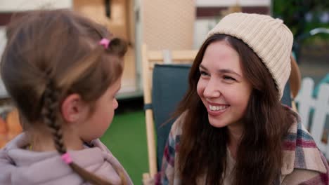 Close-up-shot-from-behind-the-shoulder-of-a-little-blonde-girl-in-a-pink-hoodie-talking-to-her-mother,-a-brunette-woman-who-hugs-the-girl-and-calms-her-down-during-their-picnic-near-a-trailer-in-a-summer-camp-outside-the-city