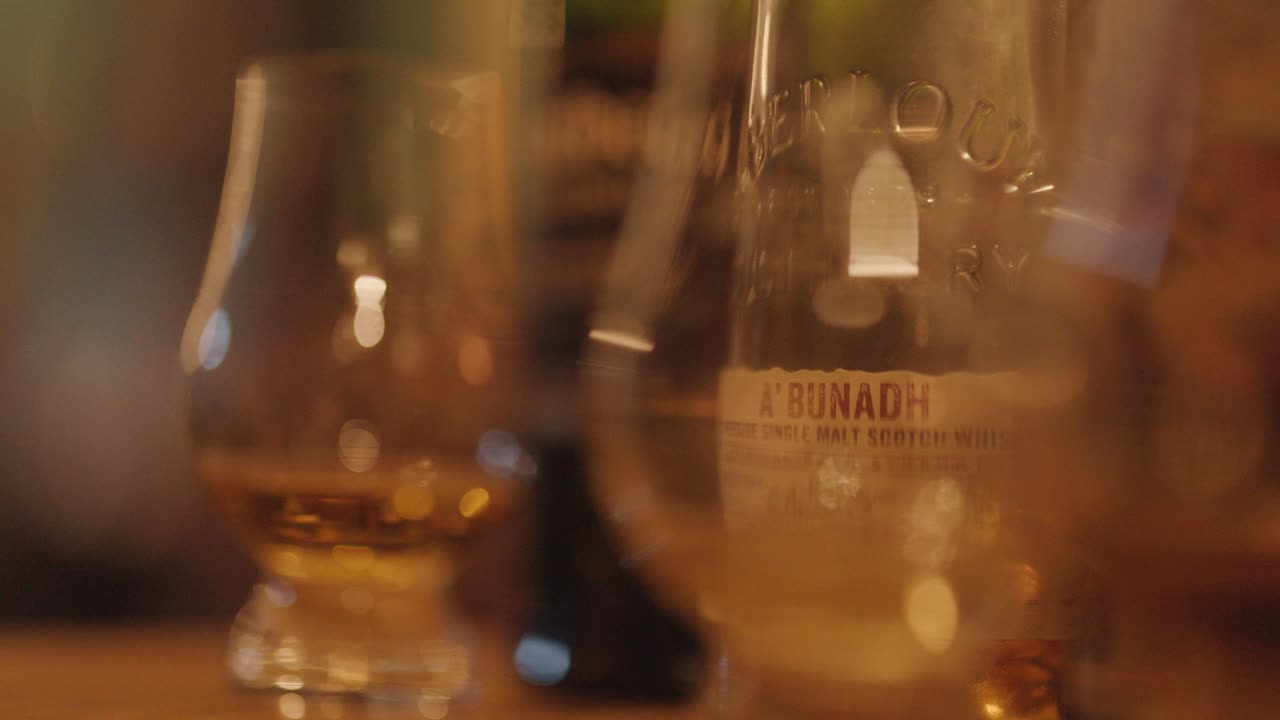 Warmly lit close-up of whiskey glasses on wooden bar, hand reaching to select a drink