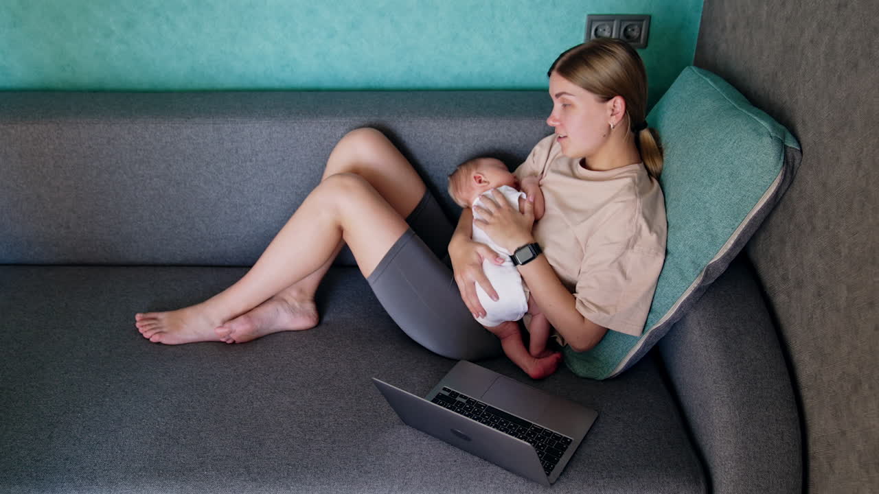 Blonde Caucasian woman with a newborn in hands sits on sofa. Opened laptop is near the lady working from home. Top view.