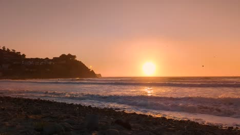 Waves-hitting-the-rocky-shore-during-golden-sunset-with-flock-of-birds-over-the-pacific-ocean