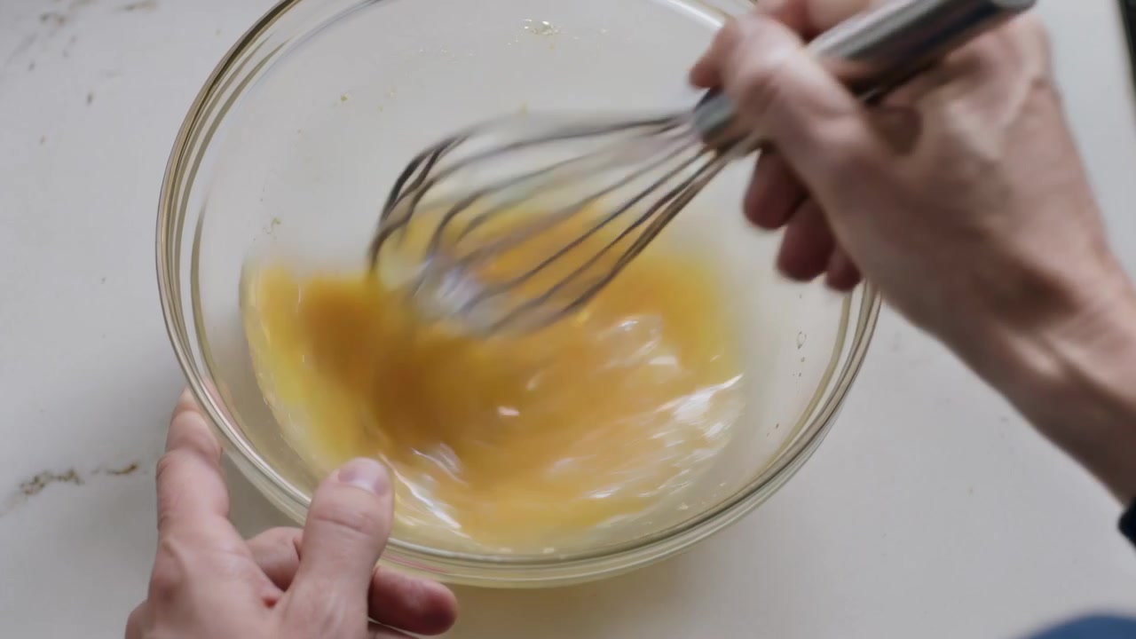 Whisking blue-cuff hands holding glass bowl, steel whisk preparing egg-mixture moving on counter