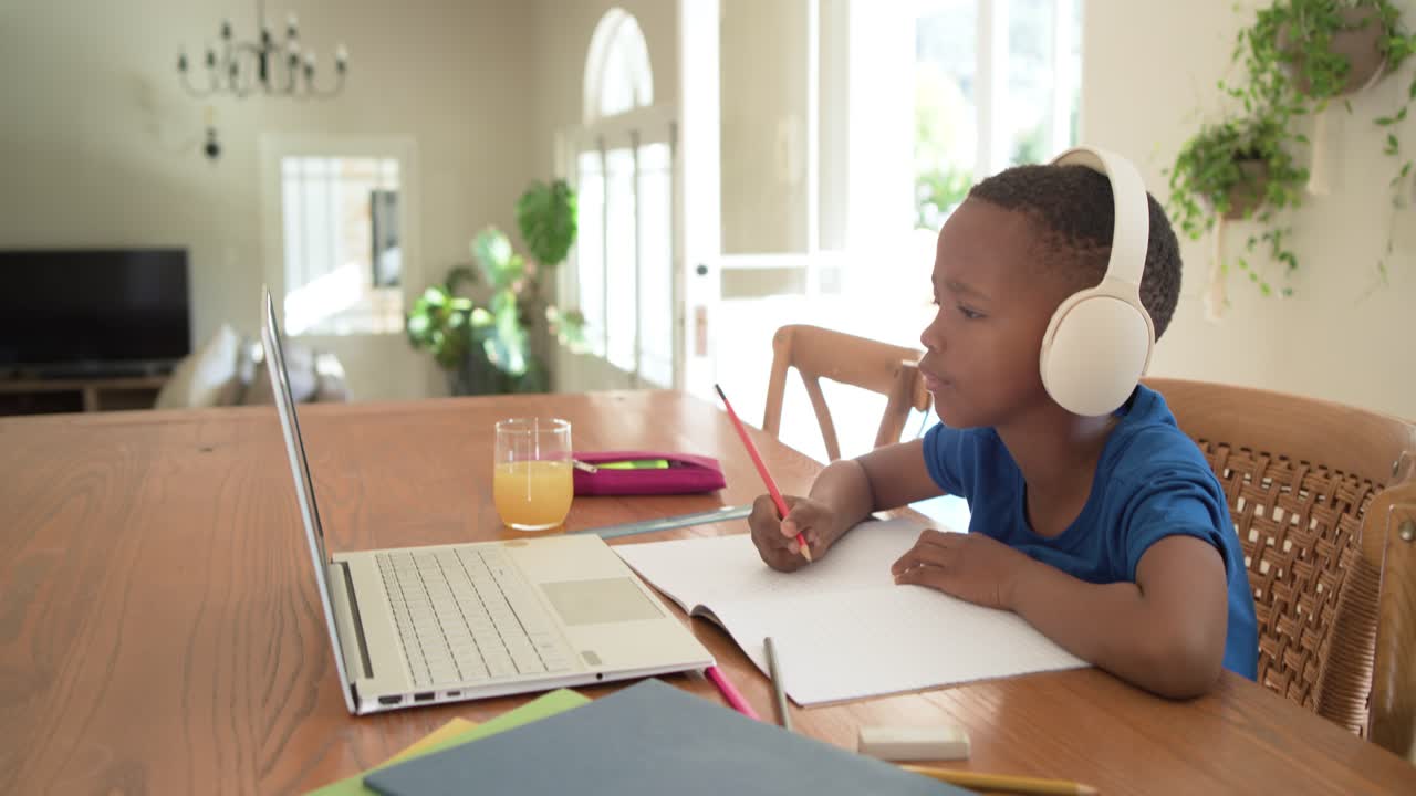 African American boy writing red pencil at dining table, raising hand to laptop prompt, copy space