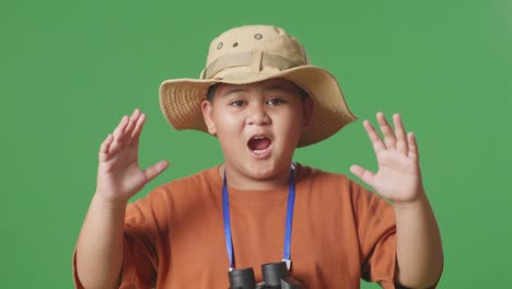 asian tourist boy with a hat saying wow after looking through the binoculars. boy researcher examines something on the green screen background, travel tourism adventure concept, close up