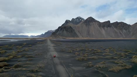 Video-De-Drones-De-La-Playa-De-Arena-Negra-Stokksnes-Vestrahorn-En-Islandia-Con-Un-Auto-Rojo-Conduciendo-Por-Un-Camino-De-Tierra