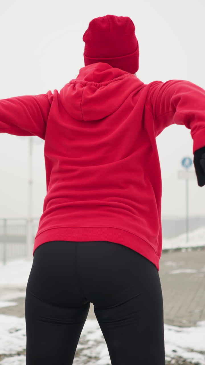 lady performing jump squats during winter outdoor workout on snowy ground wearing red hoodie and gloves beside bag on ground iron railing staircase snowy hill under foggy atmosphere breath visible
