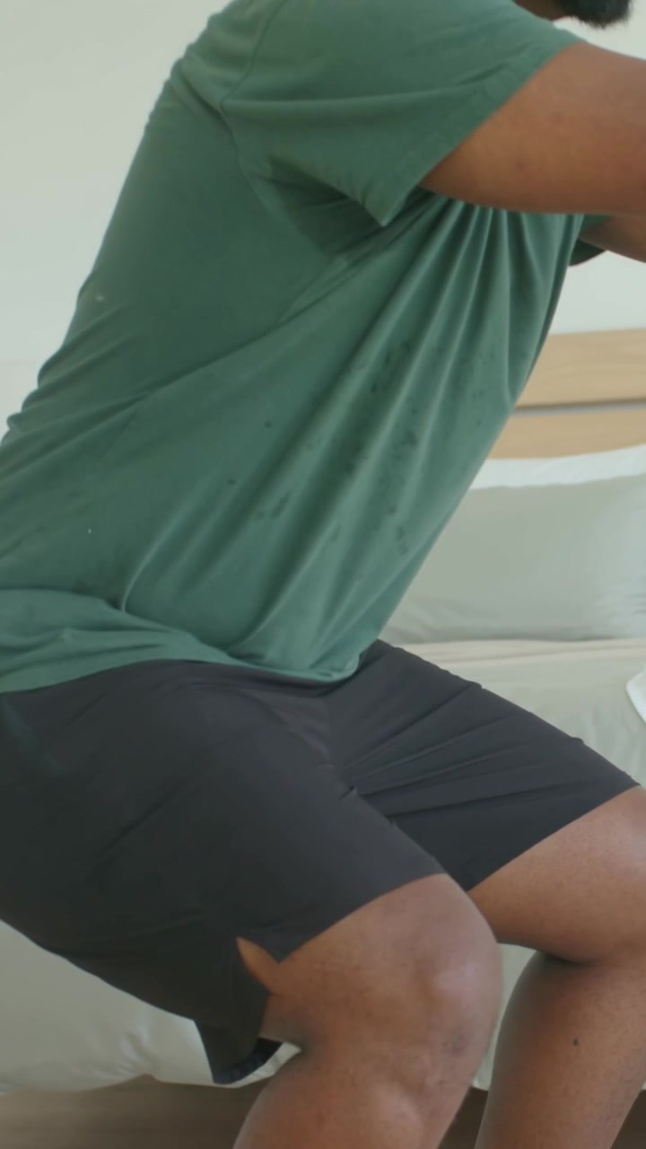 Vertical video: Leaning forward breathing hard, man in green tee squatting on bed with white watch