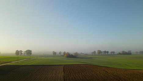 Aerial-view-above-plowed-dirt-field-and-grassy-farmland-on-hazy-blue-sky-morning