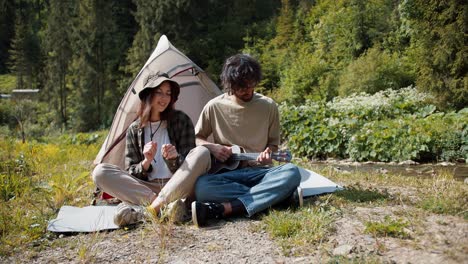 the camera zooms in on a guy and a girl of tourists who, in a special uniform for hiking, sit near the tent and play the guitar and dance near the forest