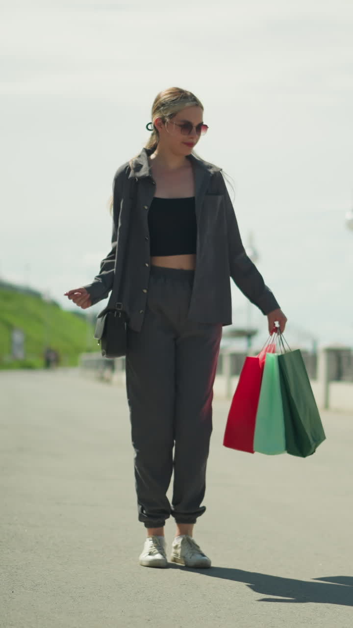 dama en camiseta negra bajo chaqueta gris baila felizmente mientras sostiene bolsas de compras en su mano derecha, su sombra es visible en el suelo con una colina a un lado y postes de luz alineando el camino