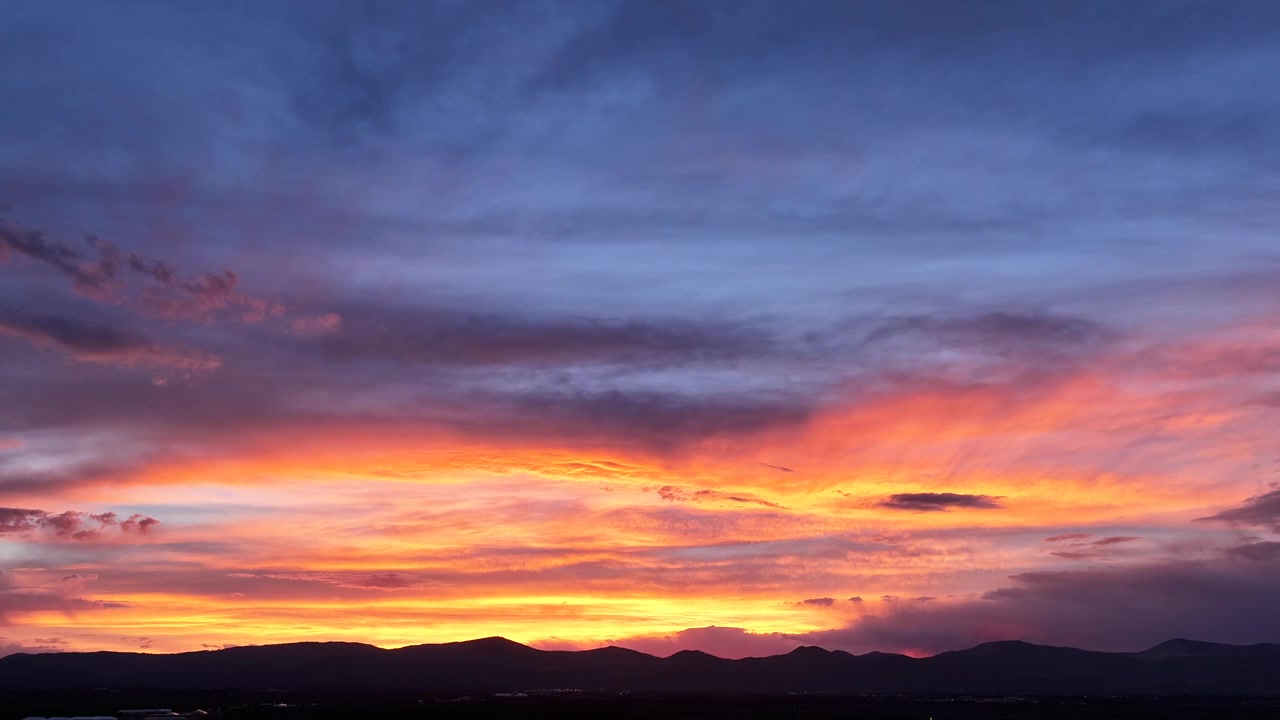En luftbåren dronetimelapse, der viser en barsk bjergkæde badet i en strålende solnedgang. Dybde, levende farver fejer hen over den skiftende aftenshimmel, mens skumringen sænker sig over de høje tinder.