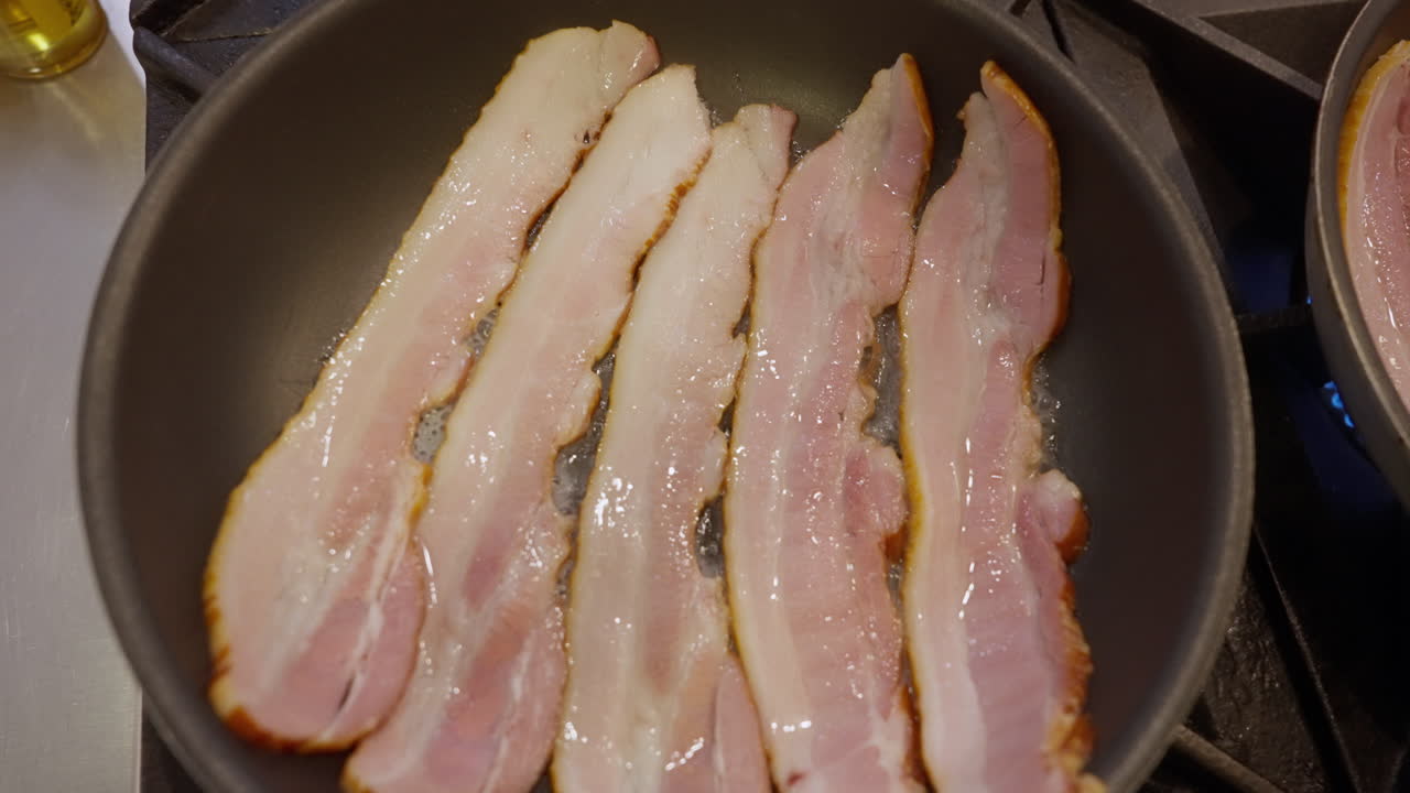 Listarelle di bacon che sfrigolano in una padella. Scena ravvicinata di preparazione con consistenza croccante che si dora, grasso bollente e vapore che si alza su un fornello: preparazione classica di cibo per la colazione in cucina.