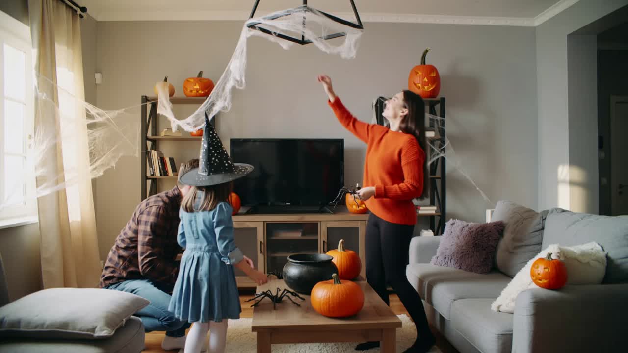 Halloween family placing spiders and hanging web in living room, daughter witch hat