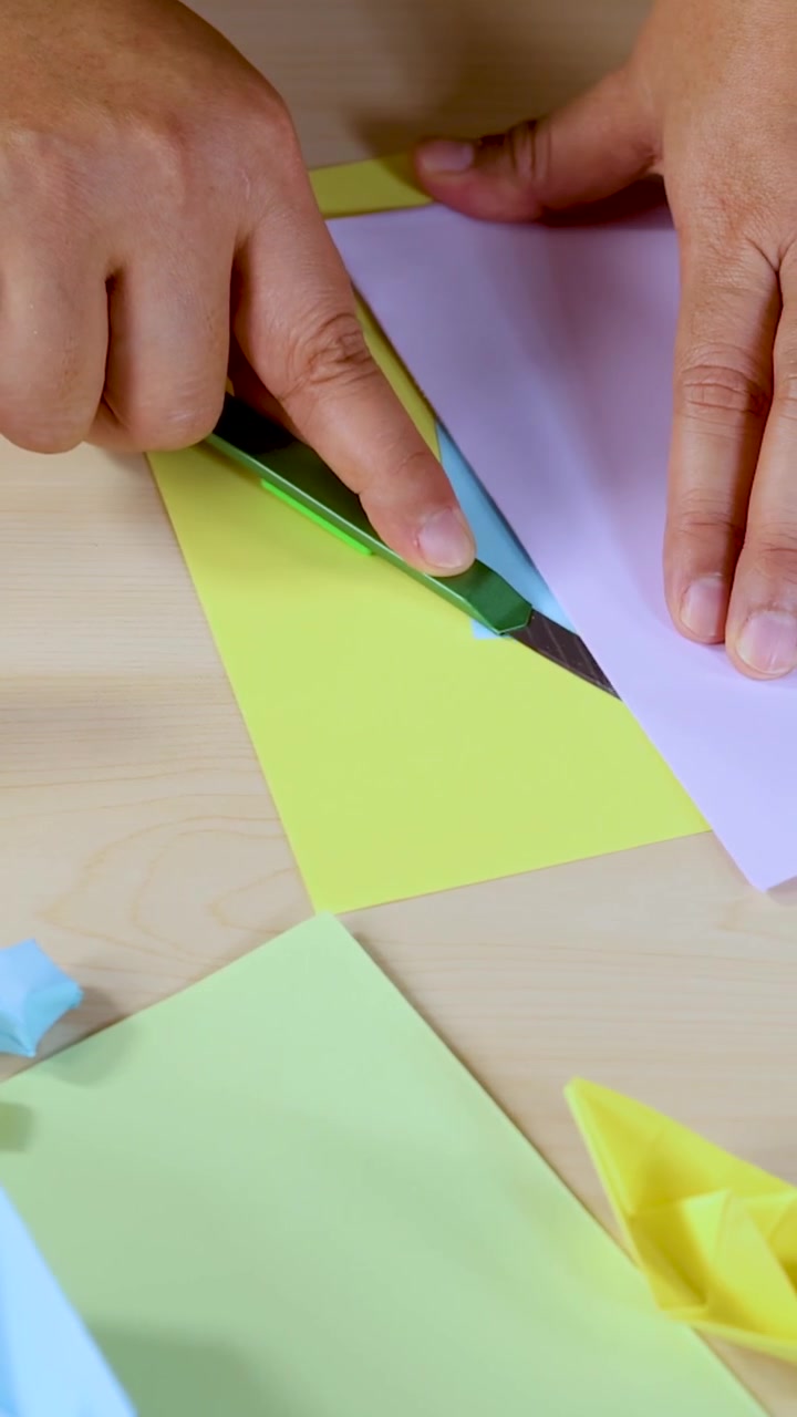 Una persona usa un taglierino su fogli pastello per origami, con illuminazione brillante e prospettiva dall'alto