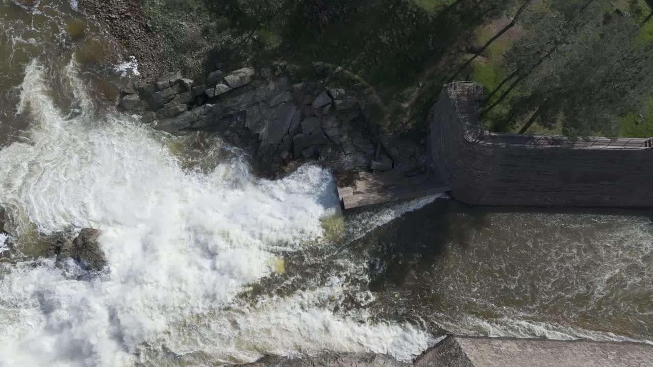 Lateral drone flight with top-down view of a dramatic spillway waterfall. White foamy water crashes into rocks, then settles and returns to the river’s course. A stunning natural transition.
