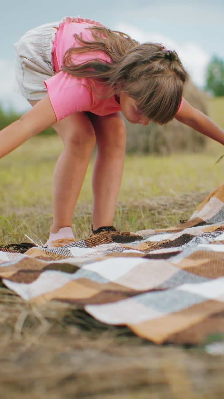 cerca de la gente extendiendo una manta a cuadros en el campo de hierba mientras el niño tira de la bala de heno en el fondo, preparación de picnic al aire libre rural bajo la cálida luz del sol con hierba seca