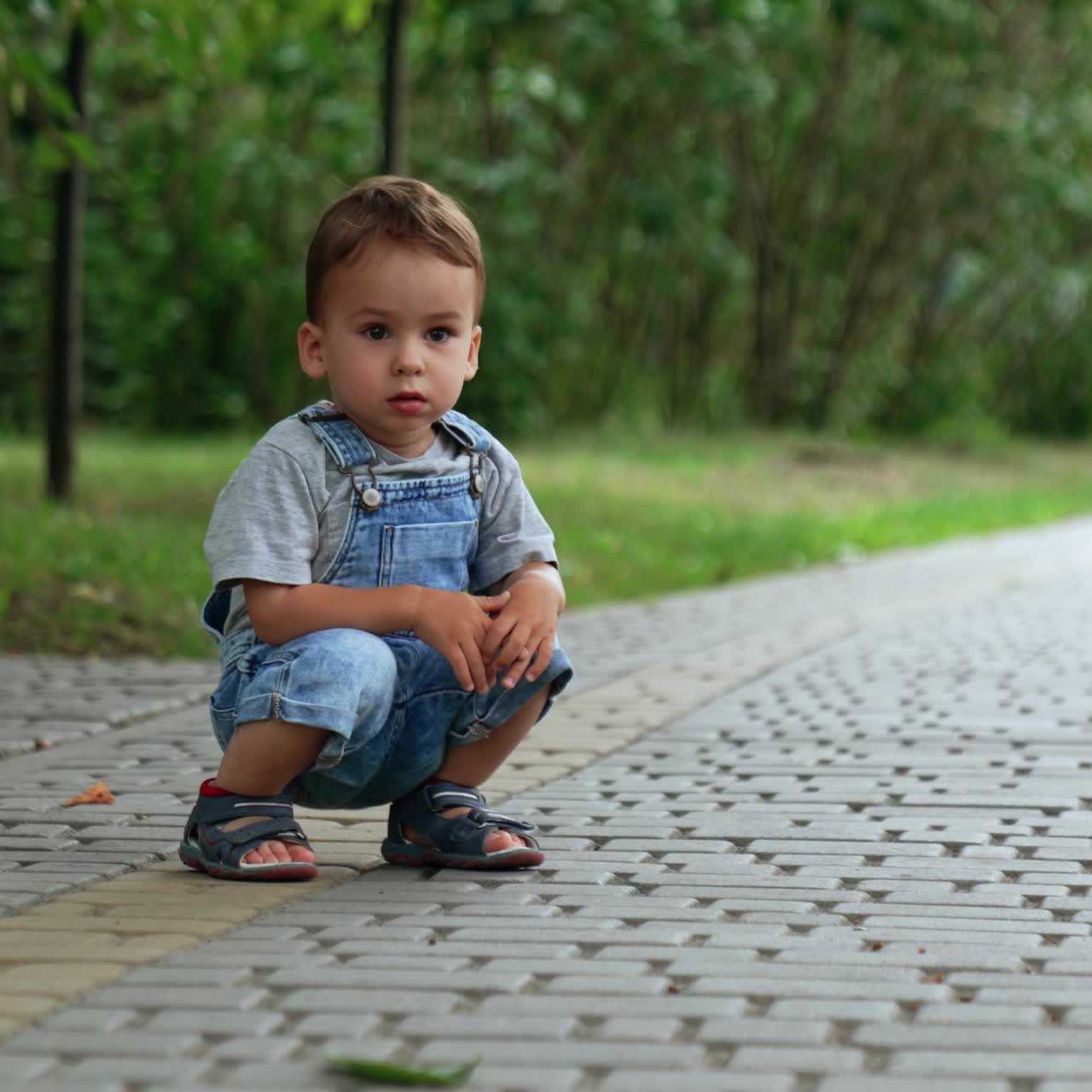 Beautiful Caucasian kid sits squatted on the paved road. Kid looks thoughtfully ahead and then runs