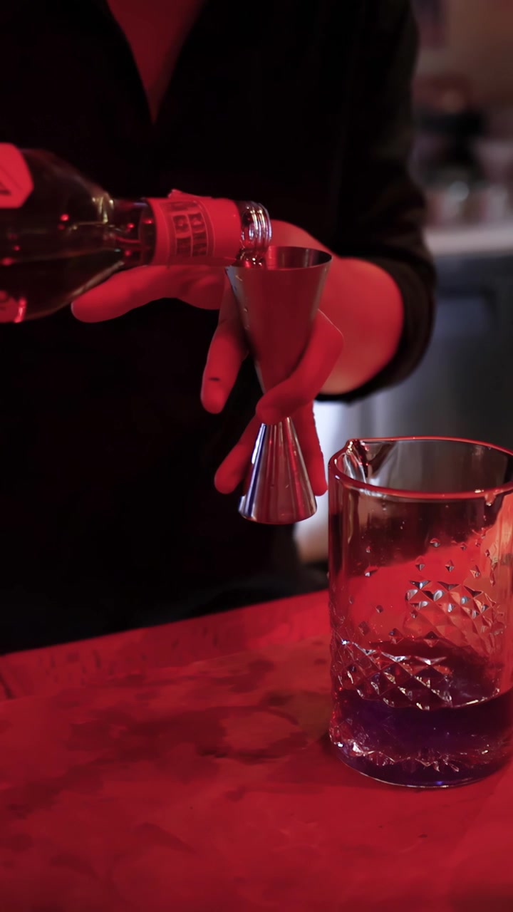 Un barman misura e versa liquore in un misurino sotto un'illuminazione rossa suggestiva al bancone di un cocktail bar.