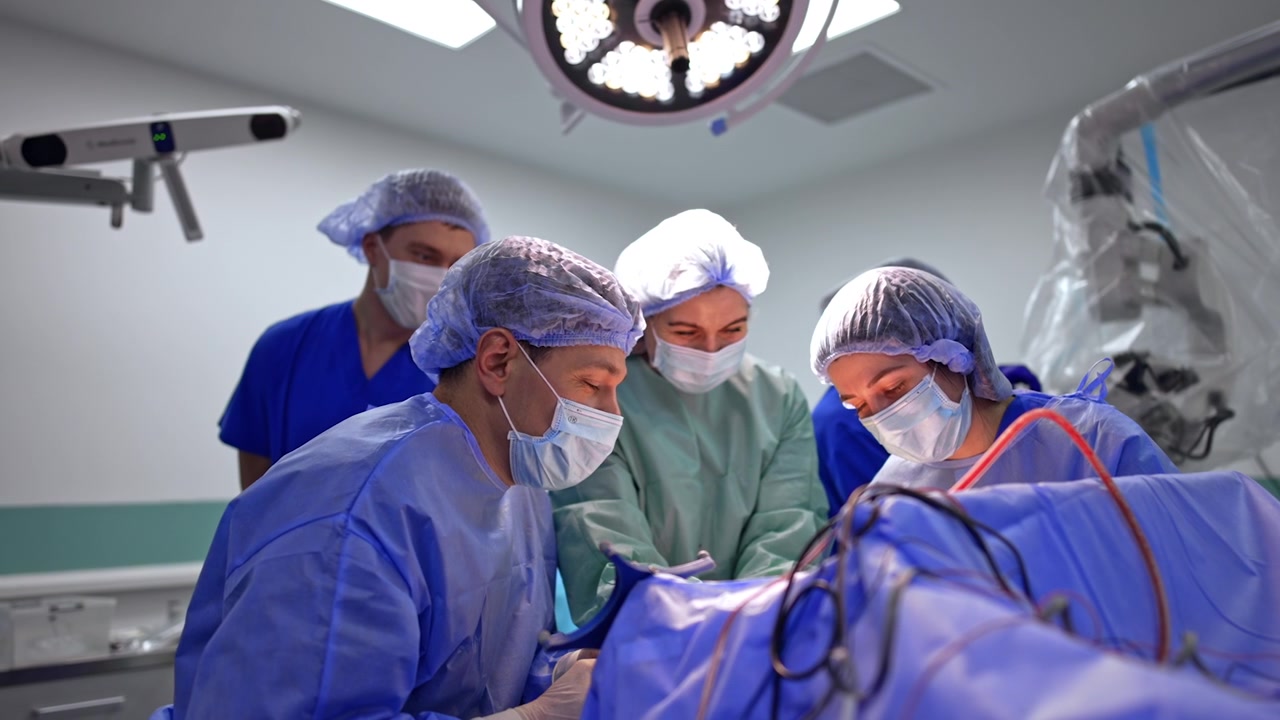Equipe chirurgica di quattro persone che esegue una neurochirurgia sotto una lampada operatoria. I chirurghi si chinano sul paziente coperto, il quarto sta dietro, con braccio di neuronavigazione e microscopio ai lati