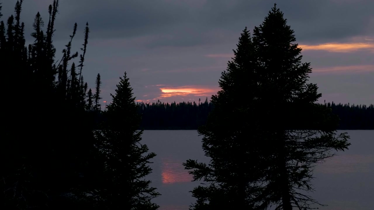 Il sole al tramonto proietta caldi riflessi su un tranquillo lago canadese incorniciato dalle silhouette della foresta boreale