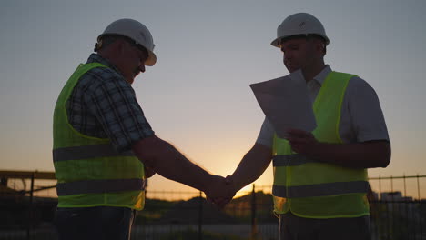 gerente de construcción y trabajadores estrechando la mano en el sitio de construcción. hombre constructor con una tableta y un hombre inspector en cascos blancos estrechan la mano al atardecer de pie. símbolo de acuerdo trabajo exitoso.