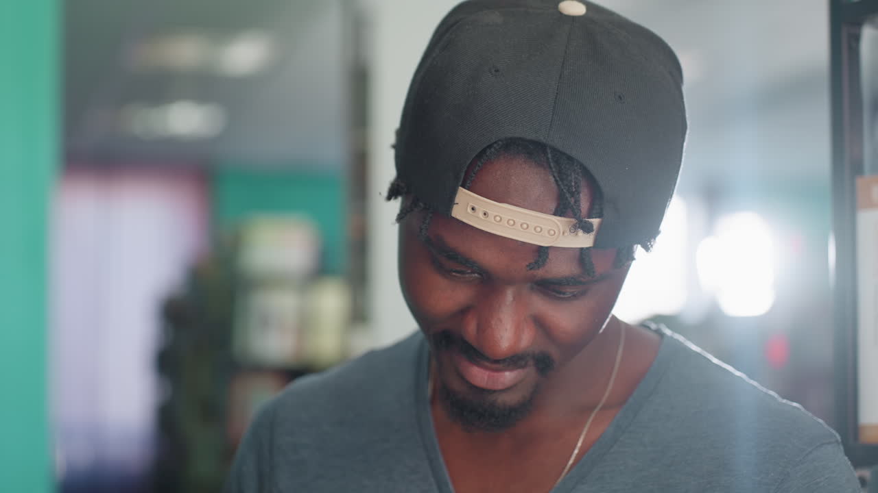 Close up of man wearing cap backward, head bowed with subtle smile, focusing intently on something out of frame, with blurred background of bookshelves and teal wall