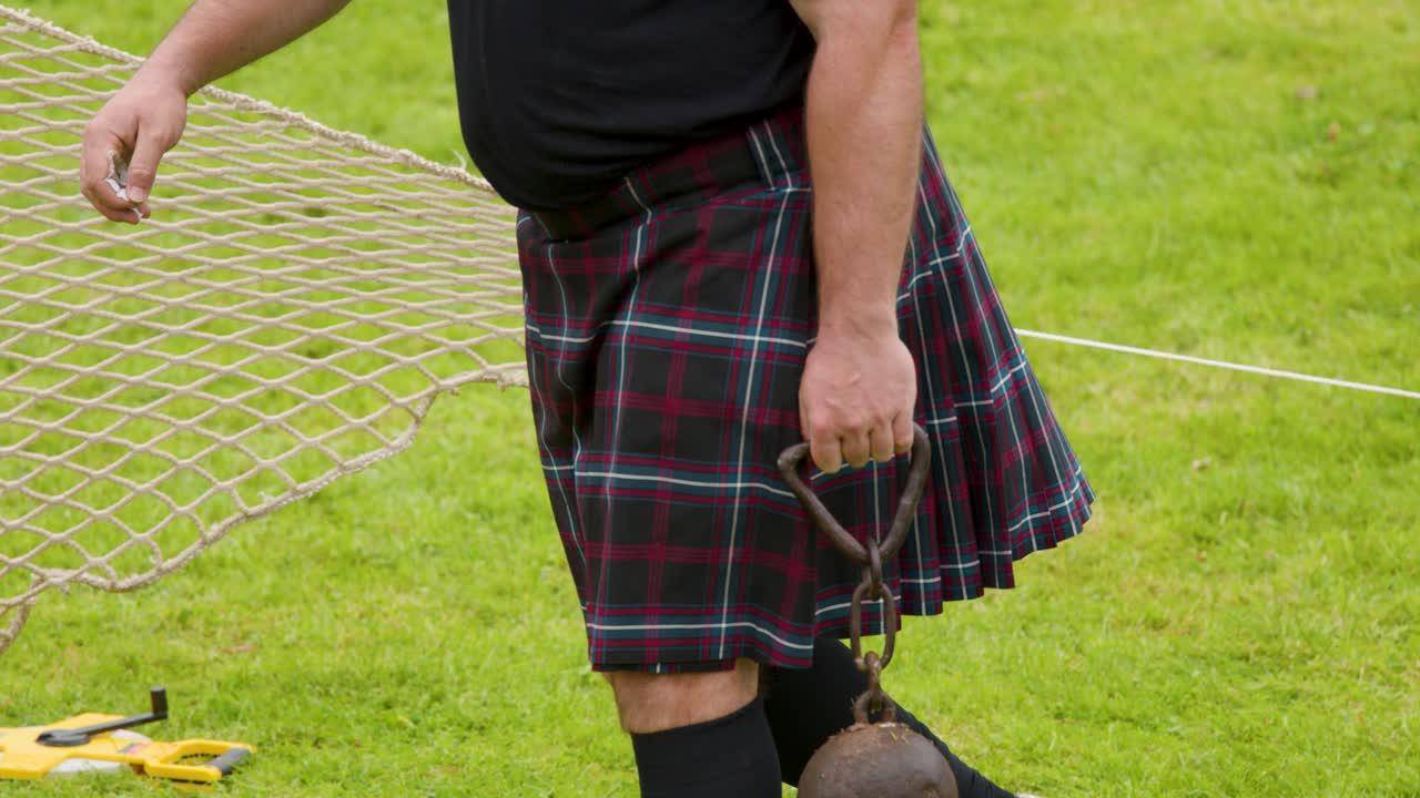 Atleta in kilt cammina sull'erba portando una pesante palla al piede durante una competizione all'aperto