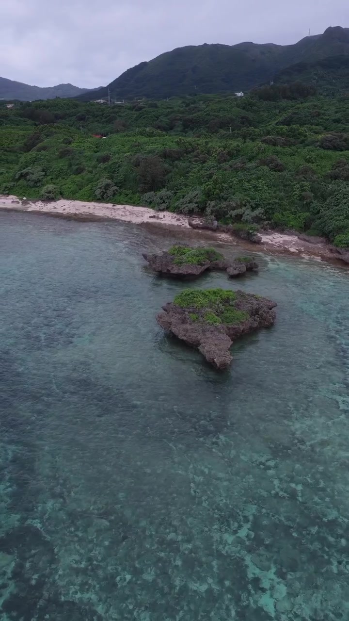 Volo aereo sopra la spiaggia di Tabaga vicino alla baia di Kabira con acqua limpida, cielo nuvoloso e nebbia bassa sulle montagne, Isola di Ishigaki, Okinawa, Giappone