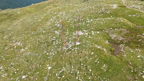 Cables-De-Alto-Voltaje-Con-Pilones-En-La-Pradera-Verde-Rocosa-De-Las-Montañas-De-Los-Alpes-En-El-Norte-De-Italia---Toma-Aérea-De-Drones