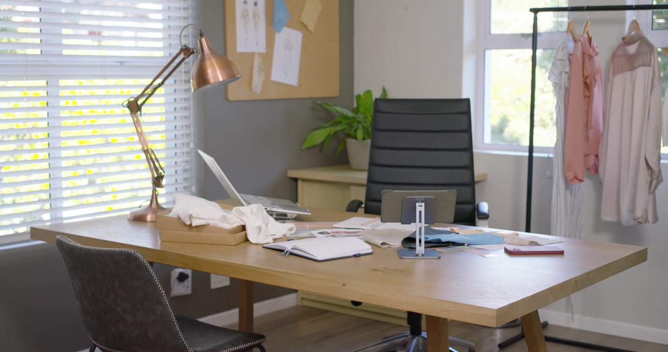 A câmera desliza para frente e para a direita, revelando uma mesa de madeira perto da janela com um laptop para revisão de design
