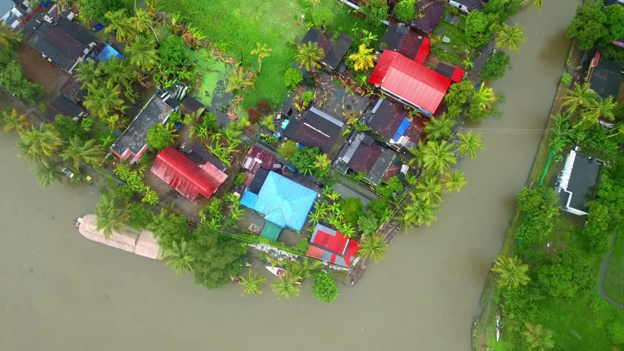Aerial top-down view of a dense tropical village with colorful roofs, squeezed between a muddy backwater canal and a bright green algae lagoon paddy field in Kerala, India