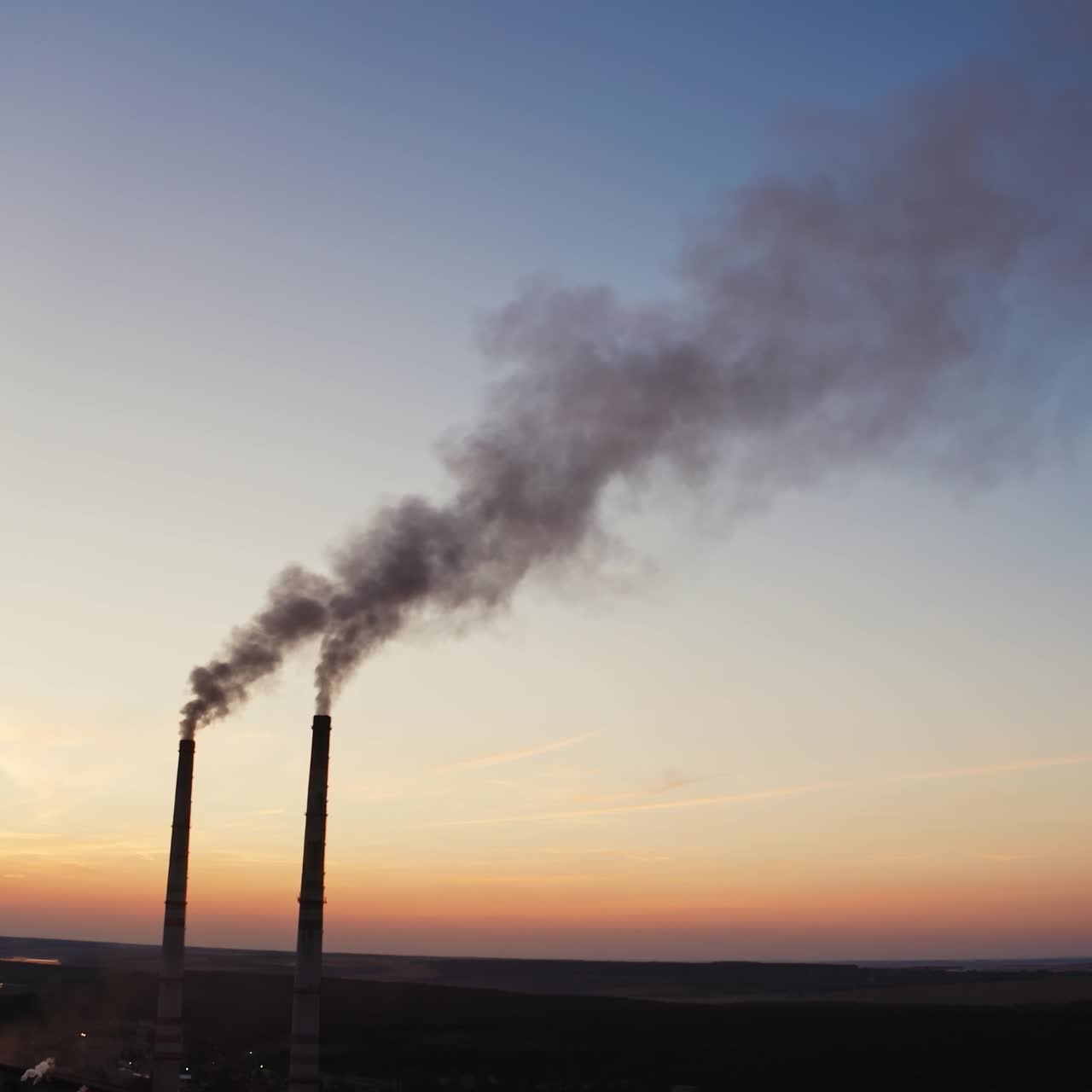 Factory with pipes among nature in the evening. Two industrial pipes with dark smoke pollute the environment at sunset.
