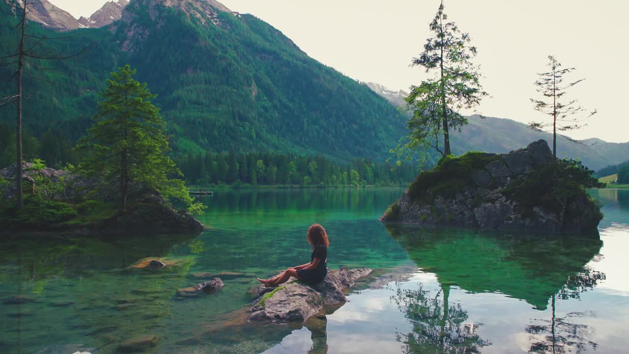 Young modern woman standing on a natural rock in the famous Lake Hintersee in Berchtesgaden, Bavaria, Germany. The scenic little island with its exceptional lush green trees in spring time. Cinemagraph / seamless video loop of the famous tourist vacation.