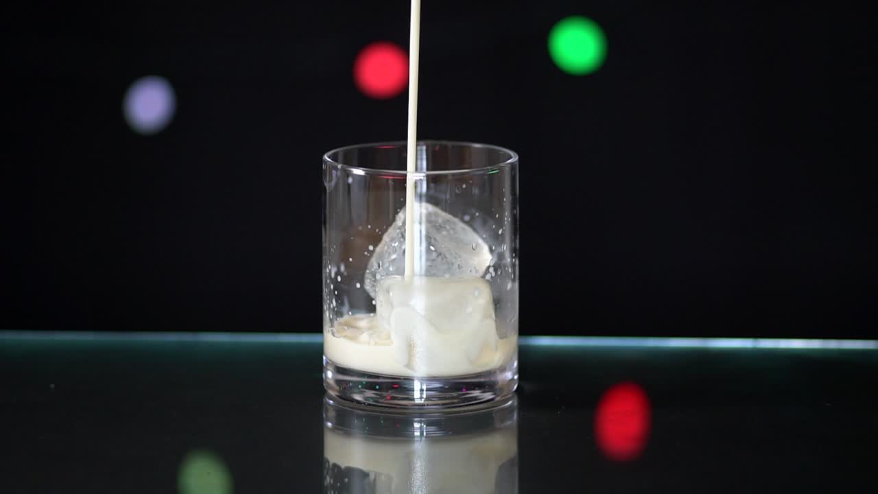 vertiendo leche de almendras en un vaso lleno de hielo, sentado sobre una superficie de vidrio con un fondo negro con luces de hadas