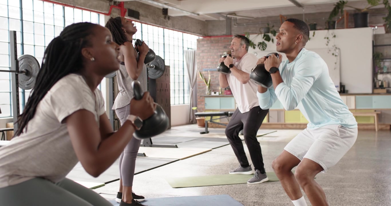 Eine diverse Gruppe von vier Erwachsenen führt auf Anweisung des Trainers Kettlebell Goblet Squats auf Matten für mehr Kraft aus.