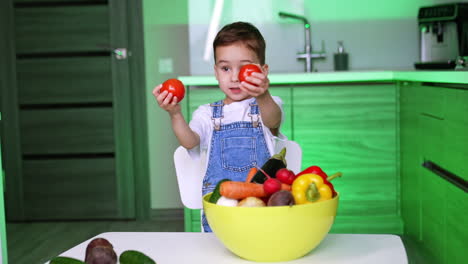 charming caucasian kid stands at the bowl with diverse vegetables. baby boy takes tomatoes showing them and putting to his eyes. child learning veggies.