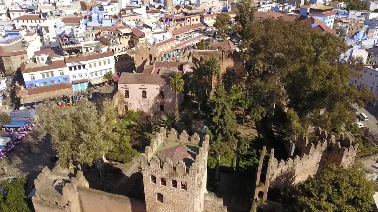 Oszałamiający, szeroki widok z lotu ptaka w HD na historyczną twierdzę Kasbah w Chefchaouen, otoczoną ikonicznymi, niebieskimi budynkami medyny. Uchwycenie unikalnego połączenia starożytnej architektury i bujnej zieleni.