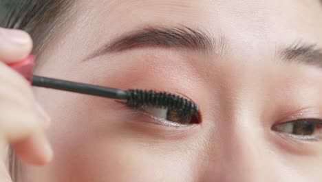 close up of beautiful young asian woman combing long black eyelashes with mascara brush and smiling while applying makeup at home