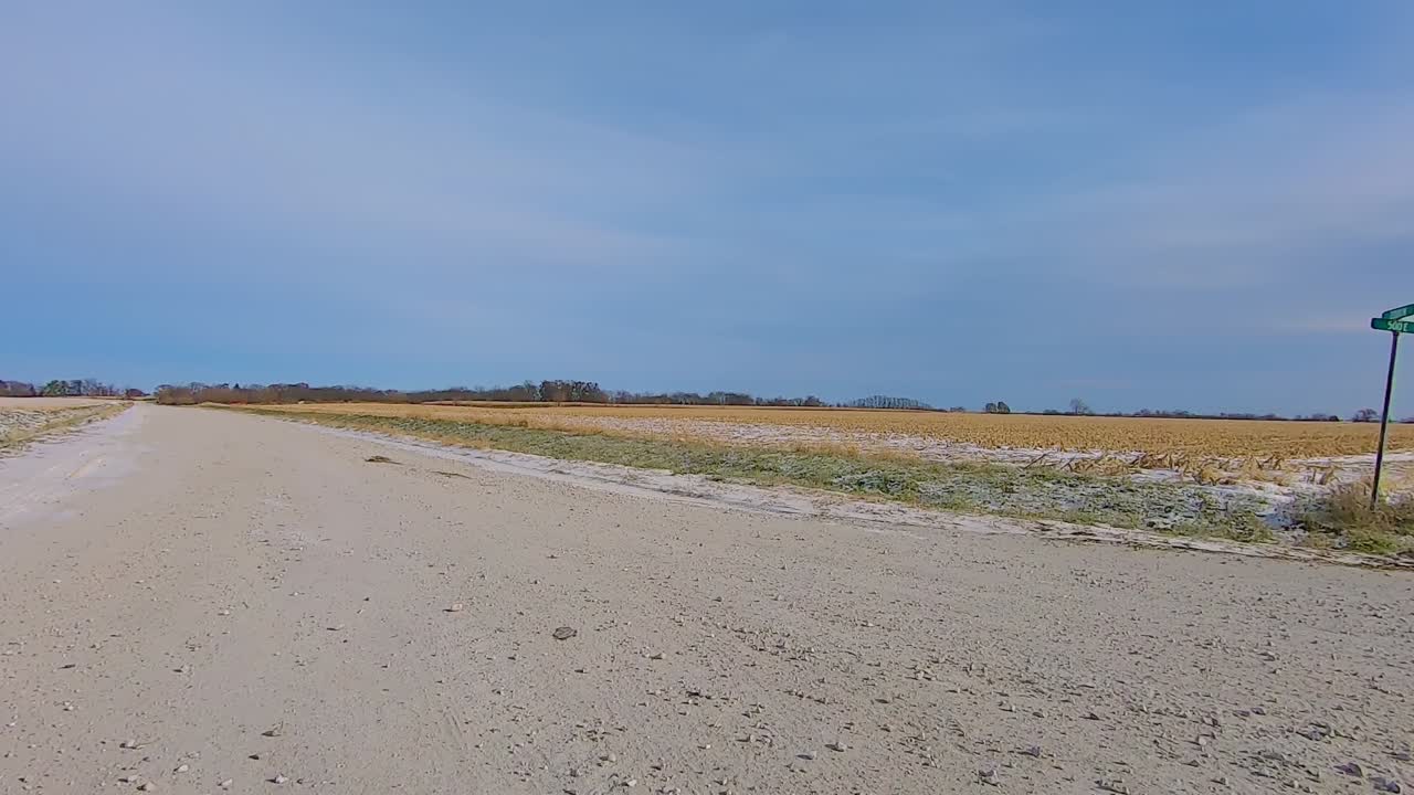 POV through the rear window while driving on a rural gravel road past empty fields on an early winter afternoon with snow in the ditch; backwards Point of View