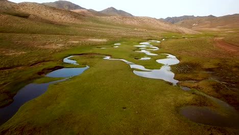 Wanderung-Inmitten-Einer-Grünen-Wiese-Mit-Ruhigem-Teich,-Bach-Spiegelt-Den-Himmel-Im-Berghochland-Im-Frühling-Wider.-Ein-Mann-Trägt-Eine-Gelbe-Gore-Tex-Regenjacke-Und-Geht-Am-Fluss-Entlang