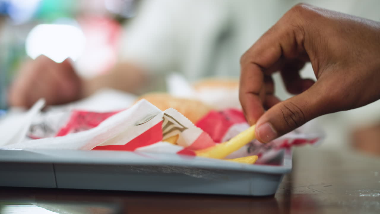 close up of hand of Indian looking man dipping into red KFC fries bag, fingers grasping crispy golden fries with oil sheen, blurred table surface behind, focus on texture and craving gesture