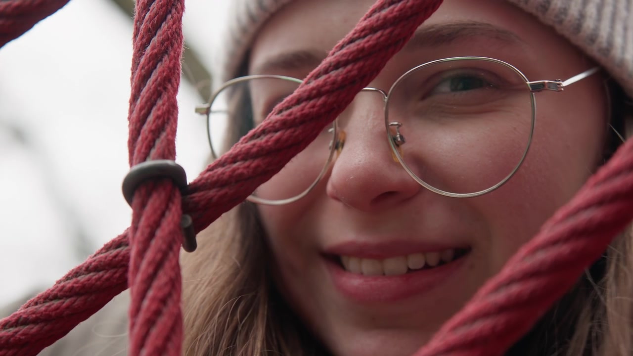 Closeup white girl kissing through rope net, winter beanie and glasses, soft smile and subtle pout, student poet pressing lips toward camera between red ropes, candid cozy portrait in overcast park