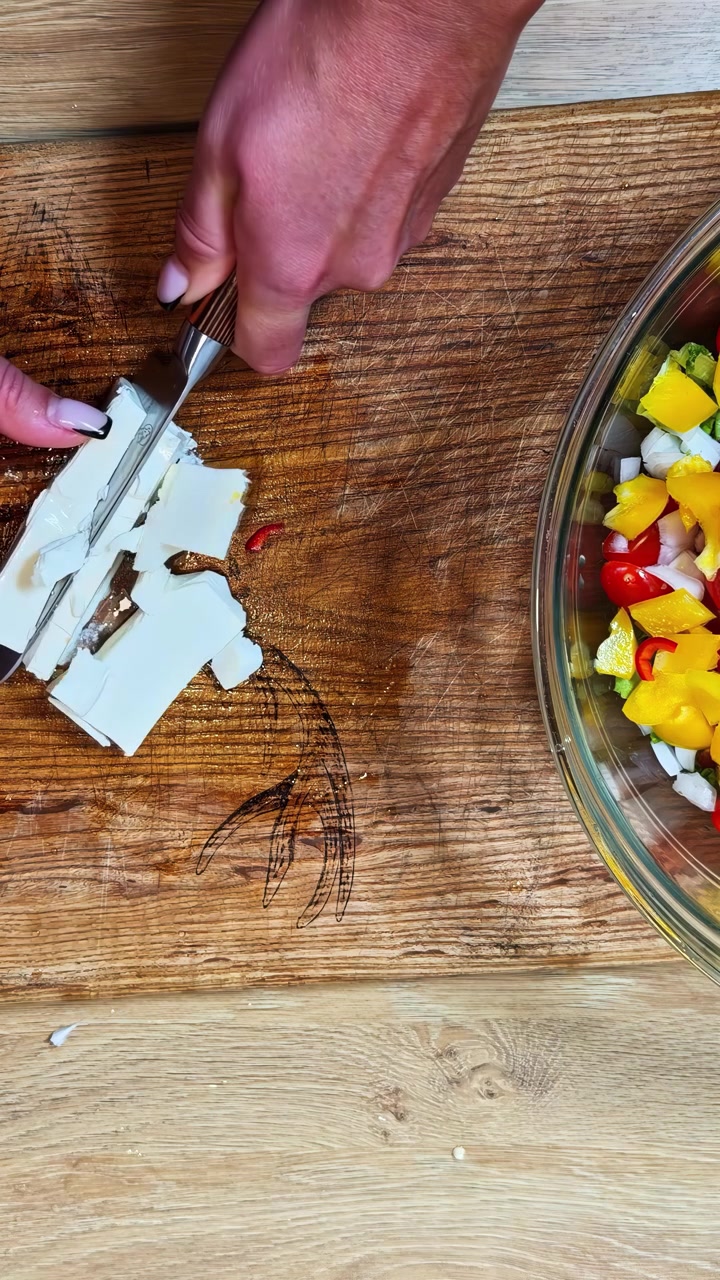 Una mano rebana queso blanco sobre una tabla de madera tallada, mientras un cuenco de cristal con ensalada colorida espera cerca, capturando una escena dinámica de preparación de comida fresca.