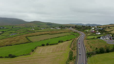 Fly-above-busy-road-winding-in-countryside-between-pastures-and-passing-through-village.-Cloudy-day-in-landscape.-Ireland