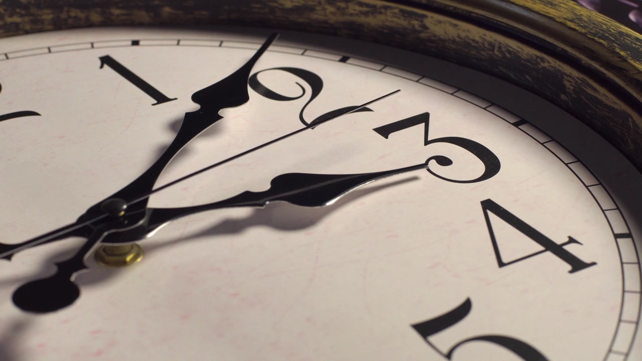 Primo piano del quadrante di un orologio analogico vintage con lancette scure e decorate. La sottile lancetta dei secondi ticchetta costantemente in senso orario oltre i numeri tre e quattro sotto un'illuminazione laterale drammatica. Scatto macro statico