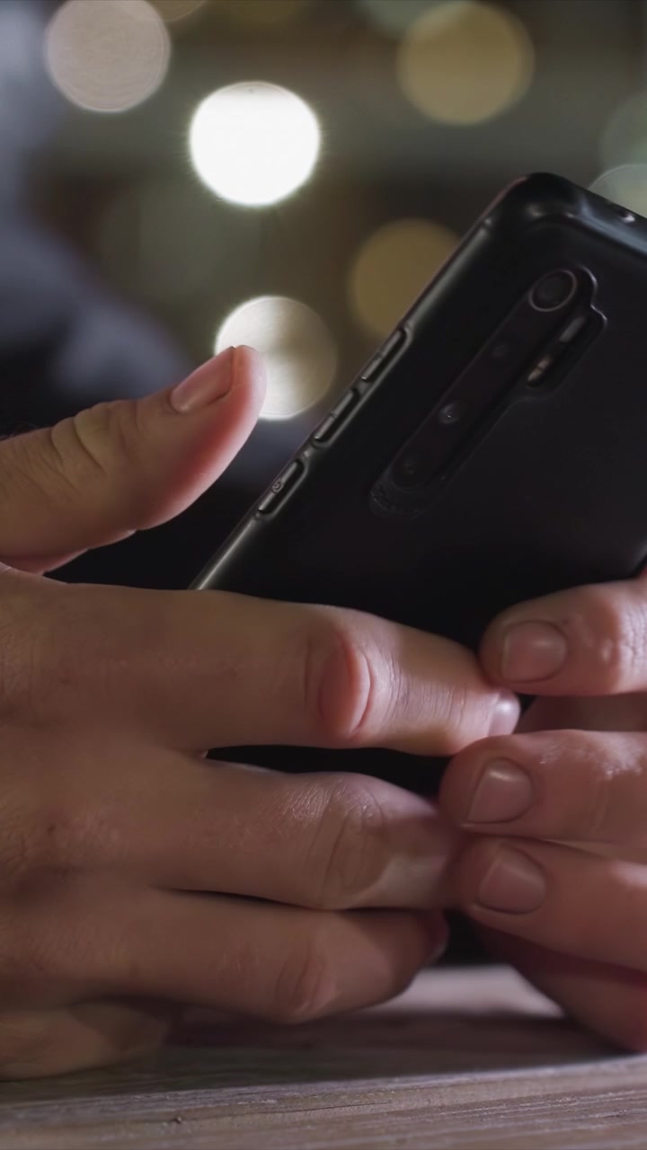 Dita stringono una custodia rettangolare per telefono in un ambiente interno poco illuminato. Il dito indice destro tocca il pulsante laterale, mentre il palmo sinistro lo sostiene. La stanza poco illuminata presenta un caldo effetto bokeh da una luce distante.